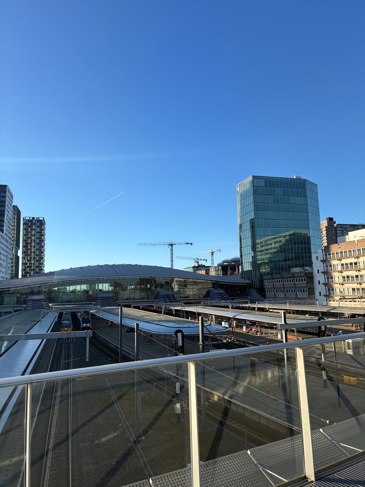 View over the platforms of Utrecht Centraal station with a train and modern glass office buildings under a clear blue sky