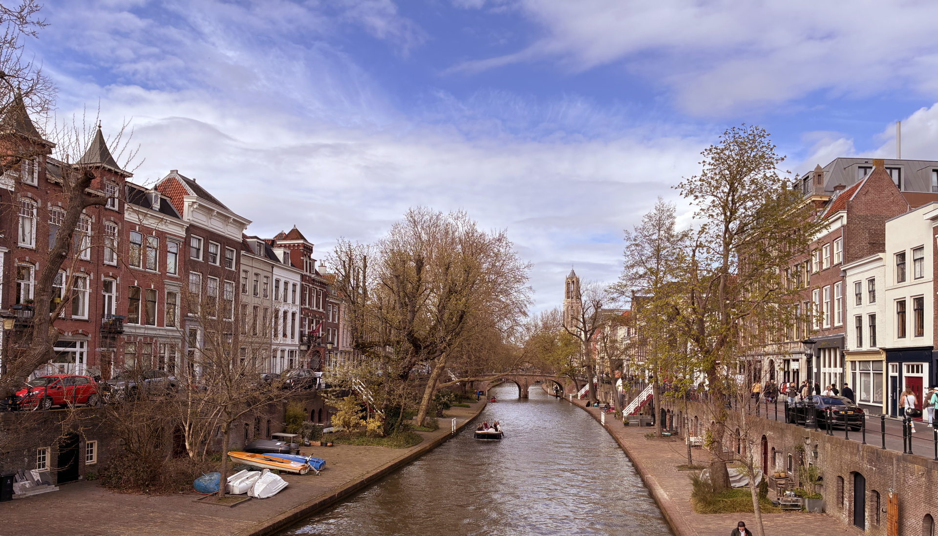 Panoramic view of Utrecht's Oudegracht canal with historic wharf houses, boats, and the Dom Tower visible in the distance under a bright blue sky